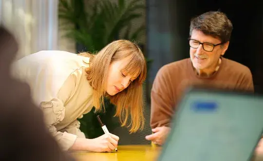 Person skriver på det gula mötesbordet och en annan sitter bredvid och ler.